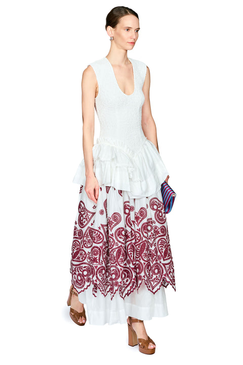 A woman modeling a sleeveless white ruffled top and a long white skirt with a burgundy paisley print, holding a striped clutch against a plain white background.