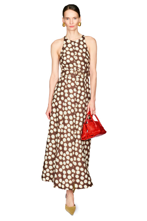 A woman modeling a sleeveless halter-neck maxi dress with a brown and beige polka dot pattern, holding a small red handbag against a plain white background.
