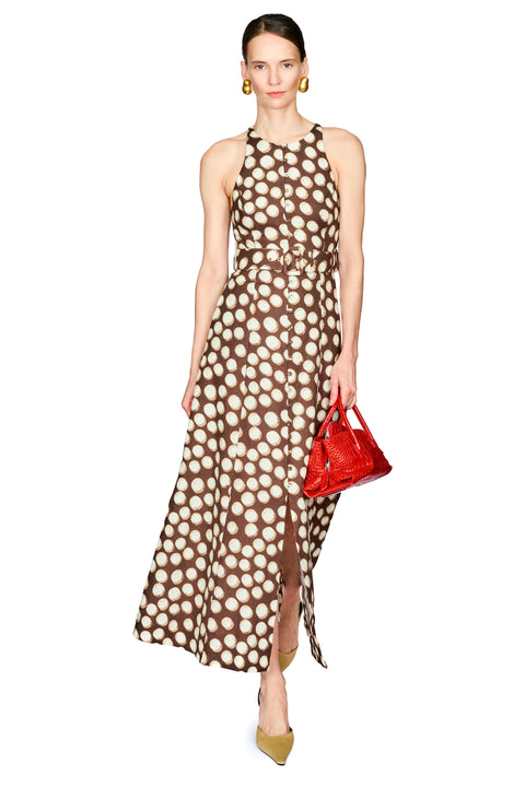 A model wearing a brown halter-neck maxi dress with a beige polka dot pattern, holding a small red handbag against a plain white background.
