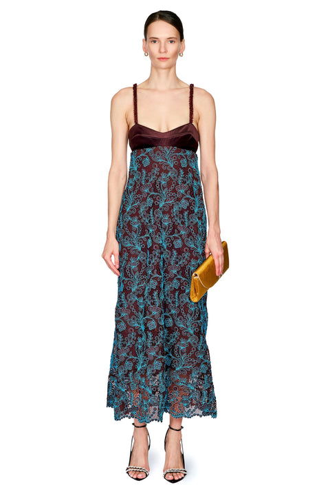 A model wearing a sleeveless maxi dress with a dark brown velvet sweetheart bodice and a teal blue floral lace skirt, holding a small yellow clutch against a white background.